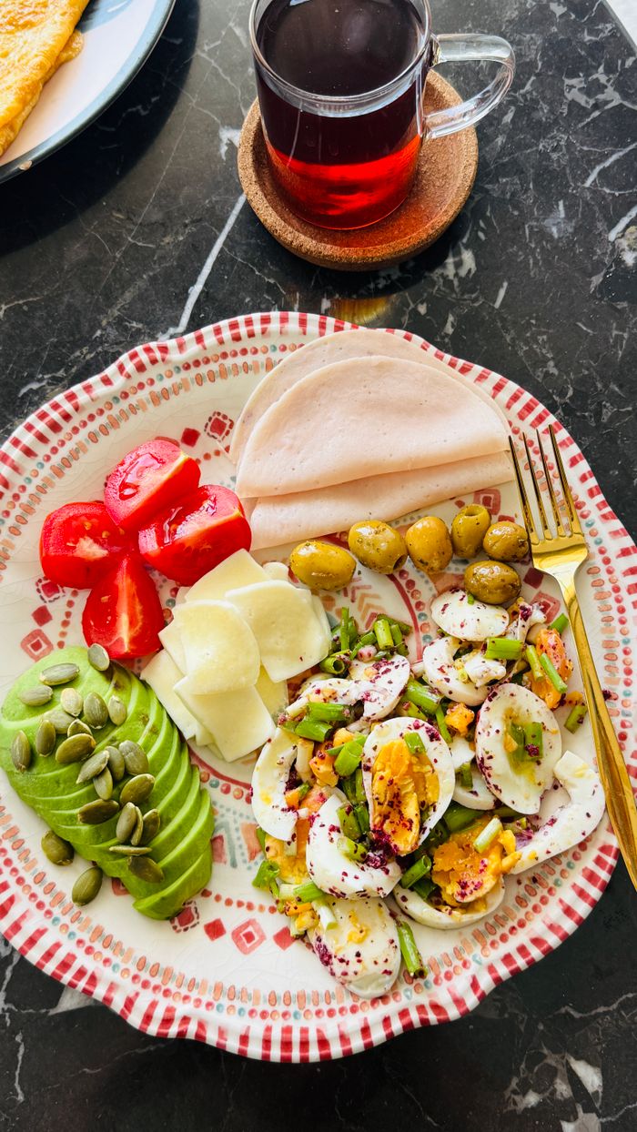 Offerings Delicious and nutritious breakfast plate featuring avocado, eggs, turkey slices, olives, and tomato with a side of tea.
