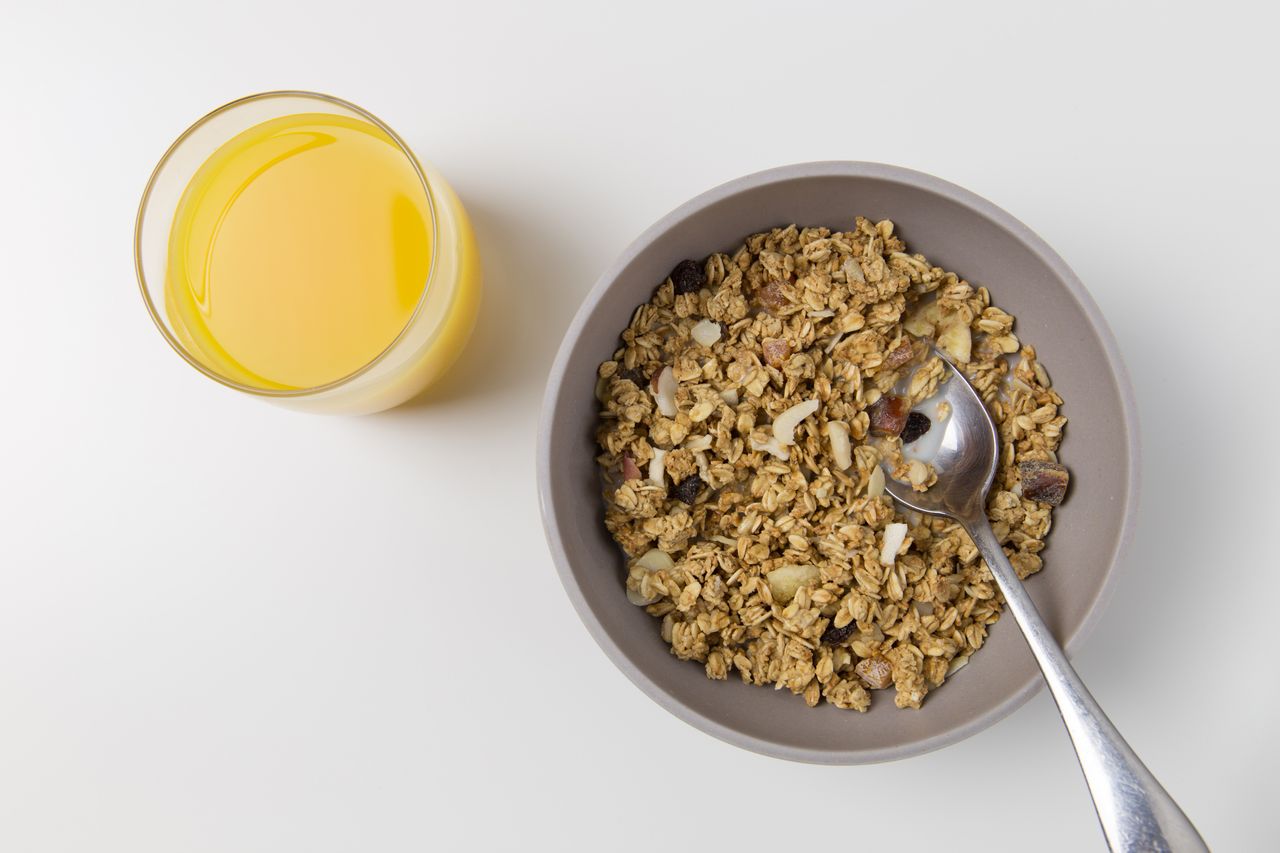 Home A top view of granola in a bowl with a spoon and a glass of orange juice on a white background.